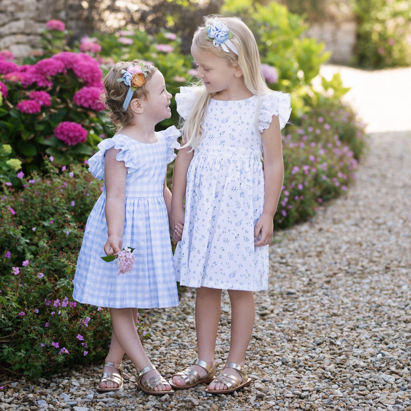 Two young girls in coordinating sister dresses standing on a gravel path with flowers and a stone wall in the background.