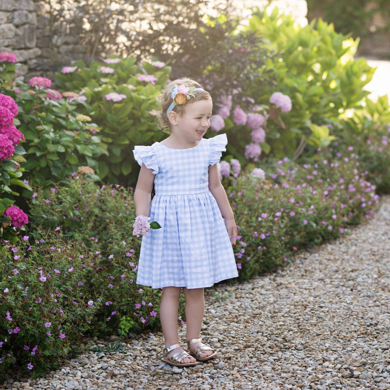 Young girl in a blue gingham dress standing in a garden with flowers and a stone wall.