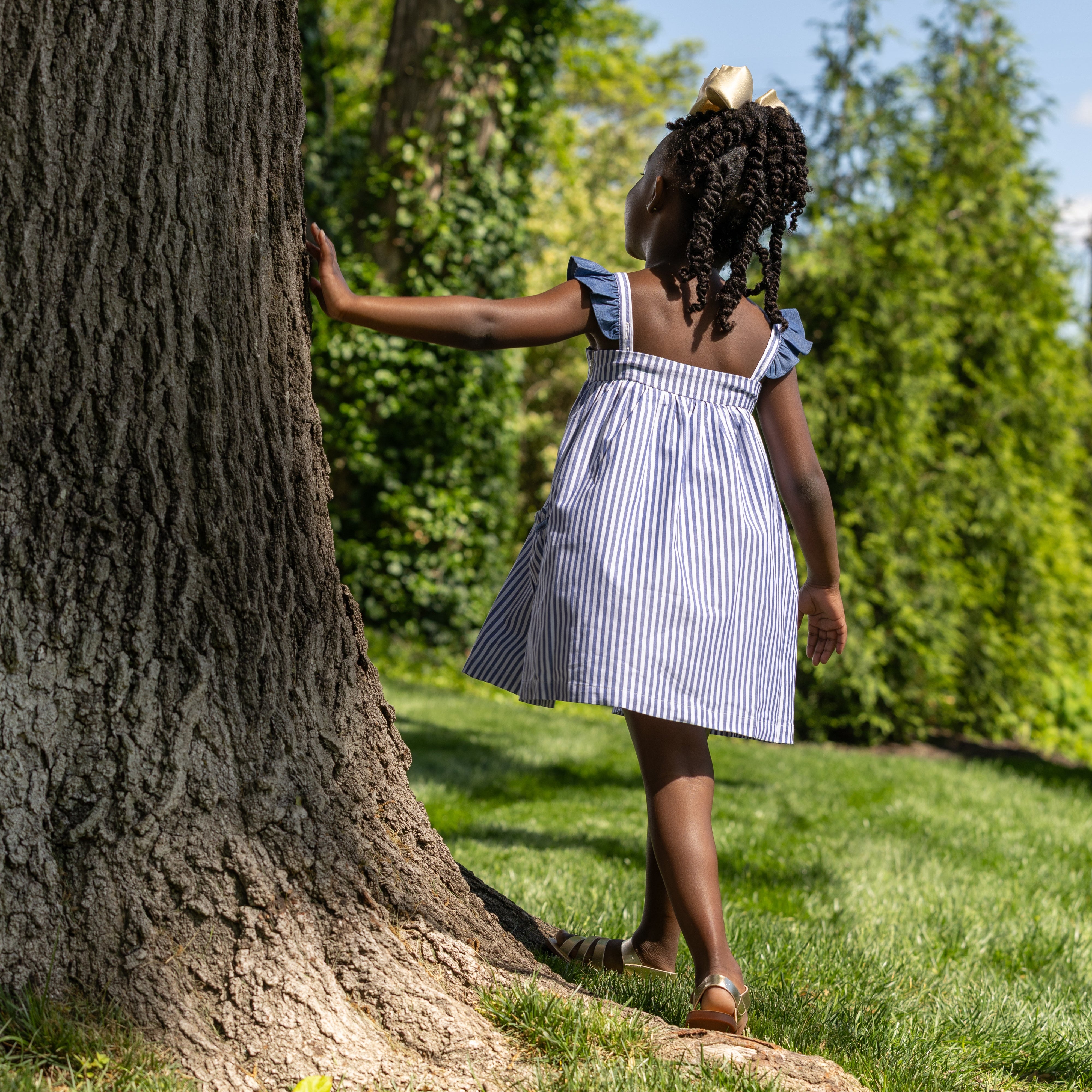 Young girl in a striped dress standing next to a large tree in a park.