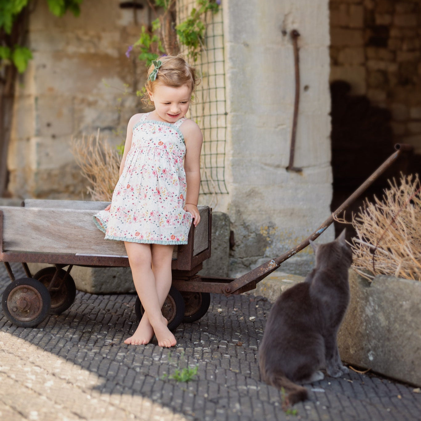 Young girl in a floral dress standing next to a cat on a stone path with a stone building in the background.
