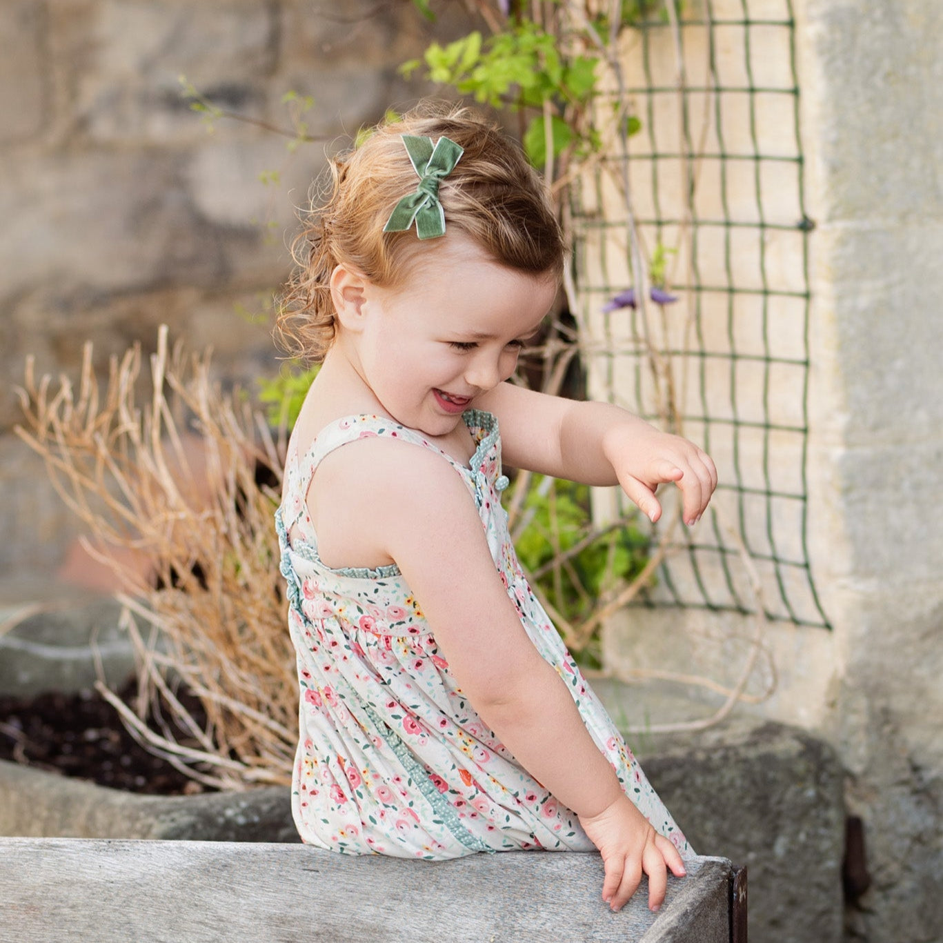 Young girl playing with a wooden box outdoors near a stone wall and plants.