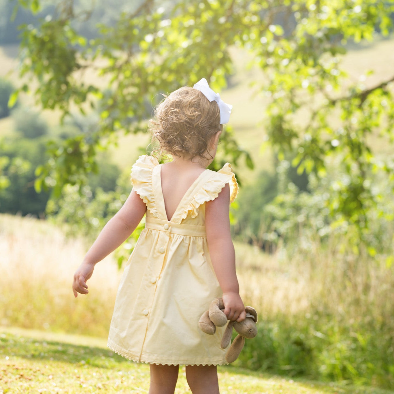 Young girl in a yellow dress holding a teddy bear, standing in a sunlit field with trees.