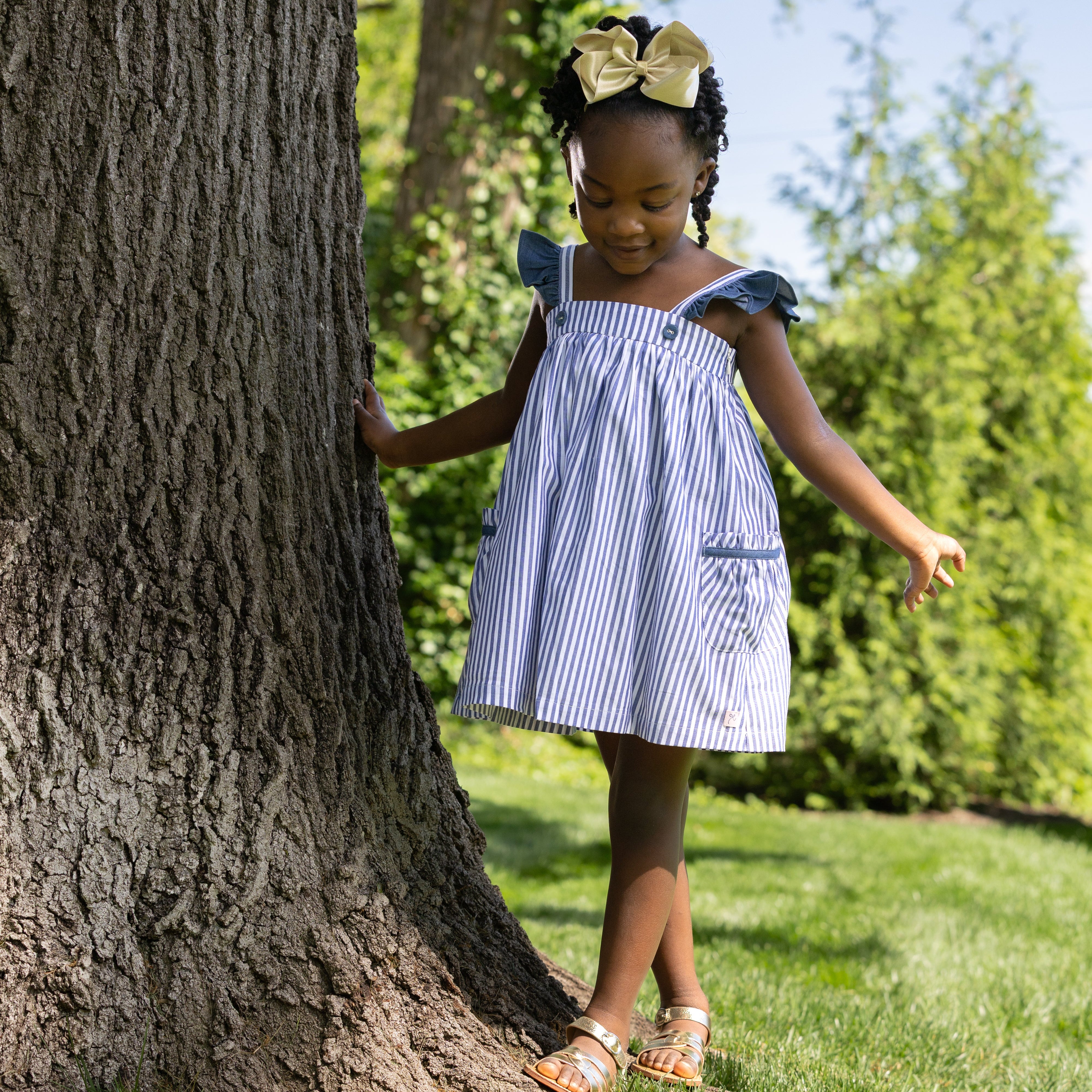 Young girl in a blue striped dress standing next to a tree in a park.